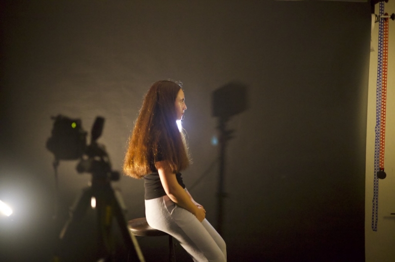 A Tisch Summer High School Photography student sits on a stool in profile posing for a portrait photo. Cameras and equipment are shown behind the student against a dark portrait background.